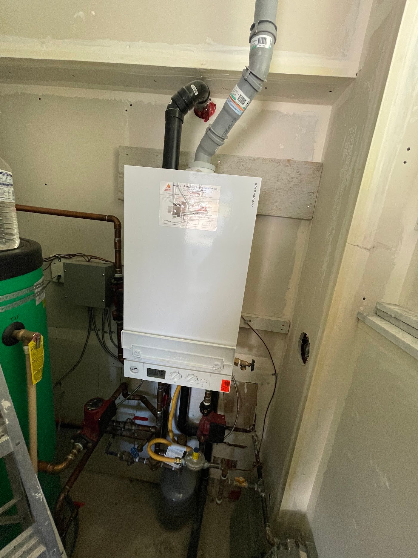 Existing boiler, adjacent tank and connected components Wall-mounted Viessmann boiler beside a green tank, with overhead vent pipes and circulation pipework below
