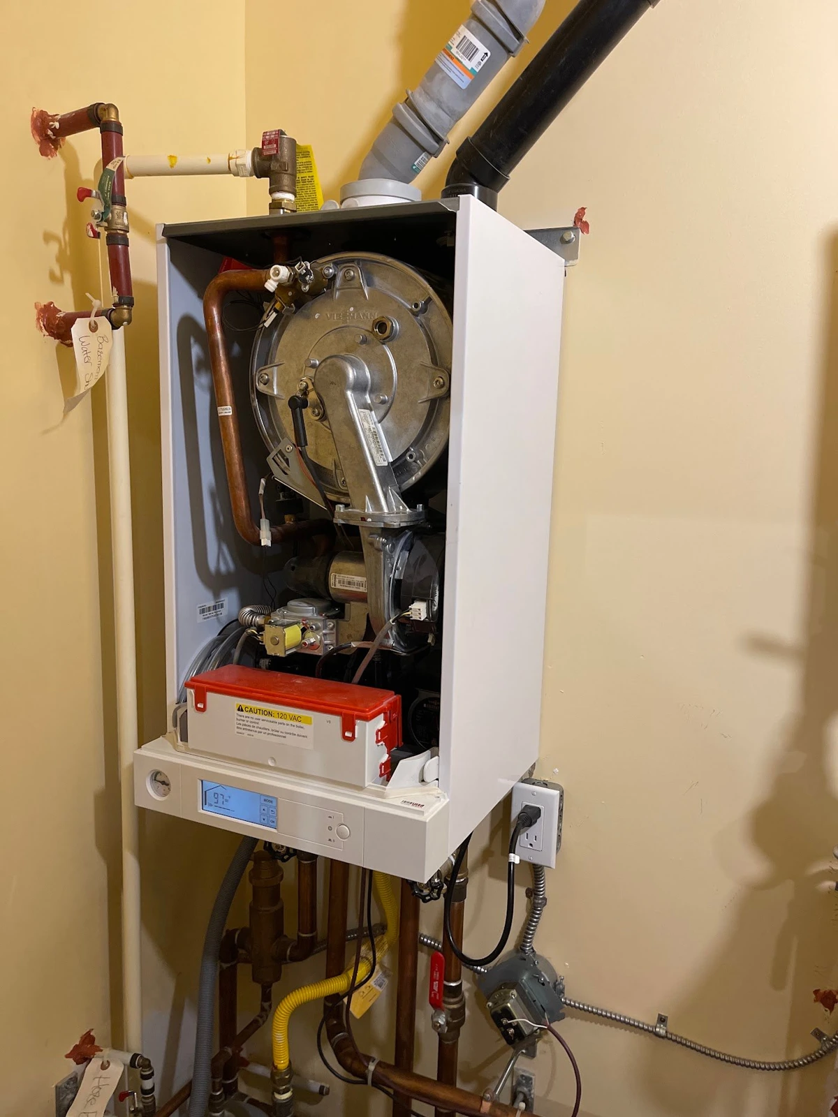 Wall-mounted boiler with its cover removed, showing internal components, vent connections and surrounding pipework.